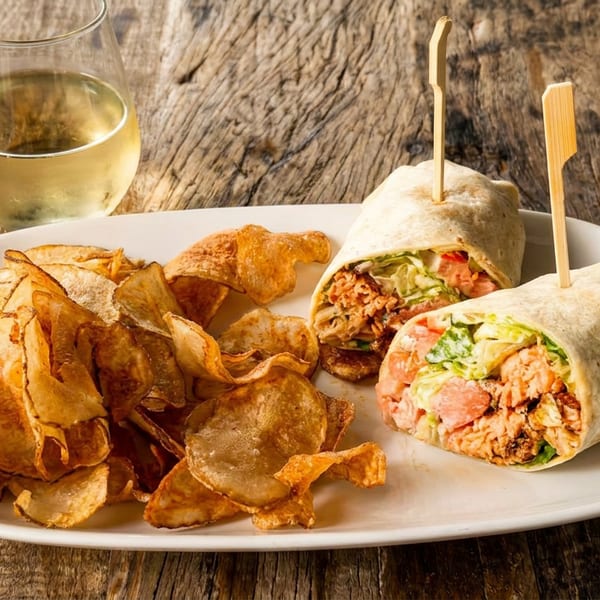 Two wraps with chicken, lettuce, and tomato, served with a side of chips and a glass of white wine.