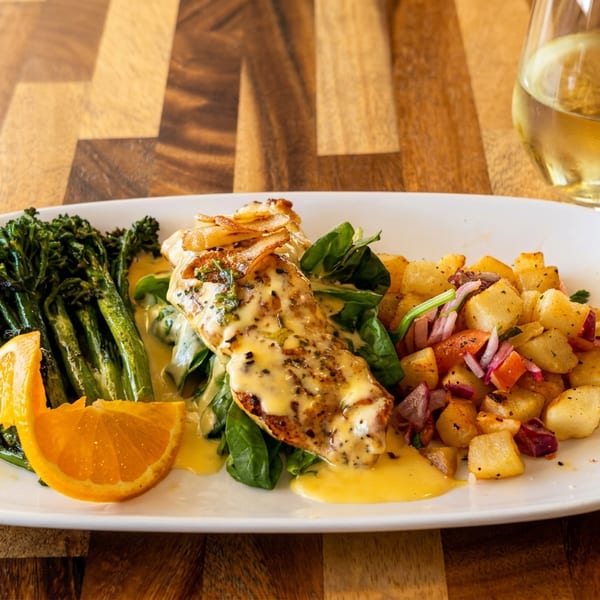 Grilled fish with creamy sauce, broccolini, roasted potatoes, and orange slices on a white plate.