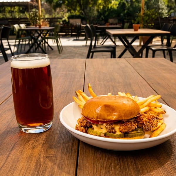 A fried chicken sandwich with fries and a beer on a wooden table at an outdoor restaurant.